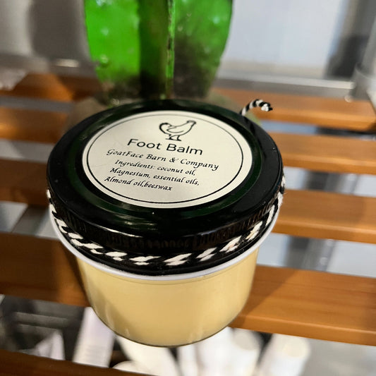 Jar of foot balm with a black lid on a wooden surface, with a plant in the background.